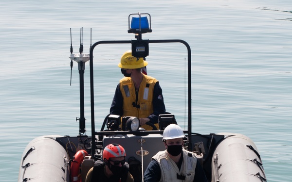 A rigid inflatable boat (RIB) with four personnel aboard is approaching or docking alongside a larger vessel. The crew members are wearing various colored hard hats and face masks, suggesting a military or coast guard operation. The boat is equipped with navigation lights, antennas, a blue emergency light, and a searchlight mounted on a console frame.
