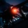Night hot refuel of PSAB F-16 Fighting Falcons during ACE capstone event at AUAB A military jet aircraft is photographed at night on what appears to be an airfield or flight deck, with a prominent orange/red navigation light illuminating the undercarriage area. The low-light, close-up composition captures the aircraft's nose section, landing gear, and underside in dramatic silhouette against a dark blue twilight sky. Colorful runway or deck lighting dots are visible in the background.