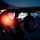 Night hot refuel of PSAB F-16 Fighting Falcons during ACE capstone event at AUAB A silhouetted ground crew technician performs maintenance or pre-flight inspection on a military aircraft during nighttime operations. The scene is dramatically lit by a bright red/orange navigation or warning light on the left side of the frame, with bokeh airfield lights visible in the background. A large spherical pod — likely a targeting or sensor pod — is visible mounted under the aircraft's wing.