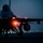 Night hot refuel of PSAB F-16 Fighting Falcons during ACE capstone event at AUAB A military fighter jet, consistent with an F-16 Fighting Falcon silhouette, is photographed on a flight line or tarmac during dusk or nighttime conditions. The aircraft is illuminated by its own navigation and position lights, with runway or taxiway lights visible in the background. A ground crew member or pilot appears to be conducting a pre-flight inspection or boarding procedure beneath the aircraft.