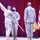 Three U.S. Army soldiers in OCP (Operational Camouflage Pattern) uniforms are participating in a formal military ceremony on a stage with a red curtain backdrop. Two soldiers appear to be exchanging or passing a unit guidon (flag on a wooden staff), which is characteristic of a change of command or activation ceremony. All three soldiers are wearing black face masks, indicating the ceremony took place during the COVID-19 pandemic period.