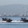 Two matching patrol boats with red trim and American flags are traveling at high speed across a bay, creating large white wakes. Personnel are visible aboard both vessels, with what appears to be mounted weaponry on at least one boat. In the background, a port facility with large container cranes, urban buildings, and tree-covered hills are visible through hazy atmospheric conditions.