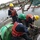 Three workers in safety gear are operating on a work boat near a waterway, handling heavy chains and rigging equipment. A partially submerged vessel or structure is visible in the background, suggesting a salvage or recovery operation. The scene appears to take place in a harbor or estuary near a residential area.