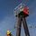 A worker in a red jacket and yellow hard hat is positioned on a service vessel alongside a navigational channel marker numbered '10', featuring a red triangular daymark. The steel pile structure includes a ladder for maintenance access and a light housing at the top. Wind turbines are faintly visible on the horizon in the background, suggesting an offshore or estuarine industrial environment.