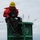 A worker in a red safety jacket and yellow hard hat is seated atop a large green navigational buoy marked with the number '9'. The individual is wearing a safety harness and holding a rope, suggesting maintenance or inspection work on the buoy. The setting appears to be on open water under an overcast sky.