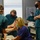 A medical training simulation is taking place in what appears to be a clinical education setting. Several healthcare professionals in scrubs and face masks are gathered around a simulation mannequin, with one practitioner appearing to practice a delivery or obstetric procedure. A 'Neonatal Resuscitation Program Reference Chart' is visible on the wall in the background, indicating this is likely an obstetric or neonatal emergency training exercise.