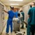 Four healthcare workers in scrubs and face masks are working in a narrow medical storage corridor lined with louvered cabinet units on both sides. A medical monitoring cart with a patient monitor screen is positioned in the center of the corridor. The staff appear to be retrieving supplies or organizing equipment in what looks like a hospital supply or equipment storage area.