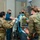 Military medical personnel in OCP (Operational Camouflage Pattern) uniforms and a civilian healthcare worker in scrubs are gathered around a patient in what appears to be a medical facility or field hospital. The team is operating medical equipment including a ZOLL EMV ventilator and IV lines, suggesting an active patient care or training scenario. All personnel are wearing face masks consistent with medical precautions.