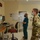 Three masked individuals — one in military camouflage (OCP/MultiCam) uniform and two in medical scrubs — are gathered in a hospital room watching a video teleconference on a wall-mounted monitor. The digital clock on the wall reads 14:24:3x, suggesting a mid-afternoon meeting. The setting appears to be a military or VA medical facility based on the visible signage and presence of uniformed personnel.