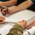 The image shows a medical or training scenario where individuals are handling ECG/EKG paper strips at a table. One person wearing military camouflage (OCP/MultiCam pattern) uniform is holding a strip of ECG readout paper, while another person holds scissors, apparently cutting the paper strip. A form or chart document is also visible on the table surface.