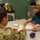 A military service member in camouflage uniform is seated at a table across from a female healthcare worker wearing teal scrubs and a black face mask. They appear to be reviewing paperwork together in what looks like a medical or clinical setting. Both individuals are wearing face masks, suggesting this was taken during the COVID-19 pandemic period.