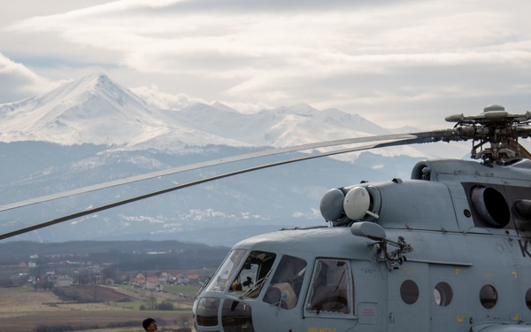 A military helicopter marked 'KFOR' (Kosovo Force) is parked on a concrete landing pad with several soldiers gathered around it. One soldier stands apart observing the aircraft while others appear to be conducting pre-flight or post-flight checks near the fuselage. Snow-capped mountains and a rural valley are visible in the background.