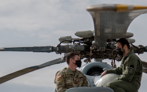 Two military personnel wearing black face masks are seated on top of a helicopter, engaged in conversation. One individual is wearing a camouflage uniform with a visible U.S. flag patch, while the other is wearing an olive/green flight suit. They appear to be conducting an inspection or discussion near the main rotor assembly of the aircraft.