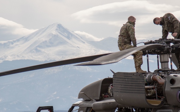 Military personnel are performing maintenance on a UH-60 Black Hawk helicopter marked with 'KFOR' (Kosovo Force), indicating a NATO peacekeeping mission. Two soldiers are working on top of the helicopter near the rotor head assembly, while others observe from ground level. The scene is set on a tarmac or airfield with snow-capped mountains visible in the background.