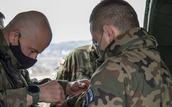 Two soldiers in Polish military camouflage uniforms are engaged in what appears to be an equipment check or adjustment, with one soldier assisting the other with gear on their wrist or arm. Both wear black face masks and display visible unit patches including what appears to be a Polish flag patch. The setting appears to be near a military vehicle or aircraft in an outdoor environment.