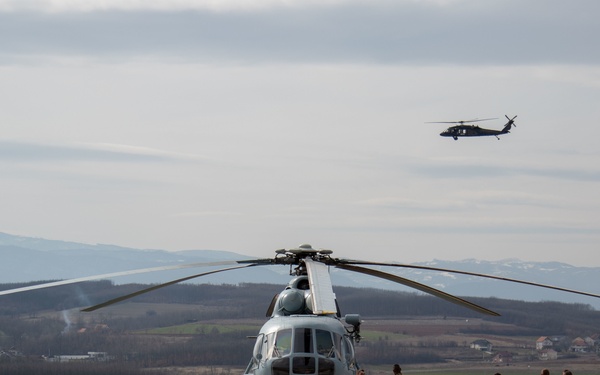 A military helicopter, appearing to be a Mi-8/Mi-17 variant, is parked on a concrete landing pad while a group of approximately six military personnel stand nearby. A second helicopter, likely a UH-60 Black Hawk, is visible in flight in the background. The scene is set in a hilly, rural landscape with snow-capped mountains visible in the distance.