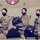 Three female U.S. Army soldiers in full OCP (Operational Camouflage Pattern) uniforms stand in front of a military helicopter marked 'United States Army'. Each soldier is wearing a black face mask and holding a flight/combat helmet. The image appears to be an official or commemorative military photograph.