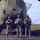 Three military personnel in camouflage uniforms and black face masks stand in front of a large military helicopter, each holding flight helmets. They appear to be posing for an official or commemorative photograph on a tarmac or airfield. The helicopter behind them appears to be a CH-47 Chinook based on its distinctive rear fuselage shape.