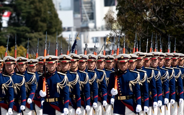 A large formation of military honor guard personnel marching in precise formation during a ceremonial parade. The soldiers are dressed in identical formal dress uniforms consisting of dark navy blue jackets with gold braiding, white trousers, white gloves, and white peaked caps with red trim. Each soldier carries a rifle with a fixed bayonet at shoulder arms position.