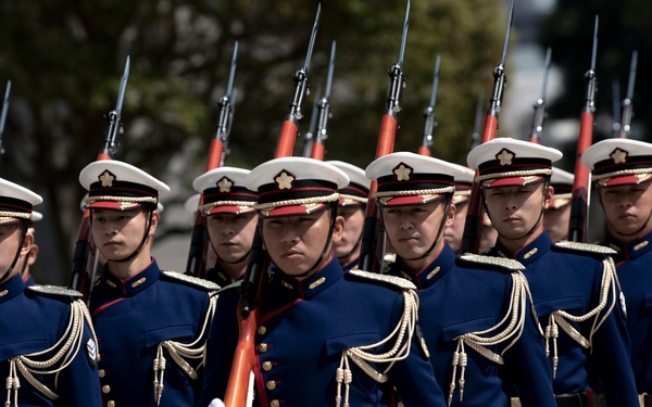 A formation of military honor guard personnel marching in precise formation during a ceremonial parade. The soldiers are dressed in formal dress uniforms featuring dark navy blue tunics with gold aiguillettes, white trousers, and white peaked caps with red bands. Each soldier carries a rifle with a fixed bayonet, held at a uniform angle.