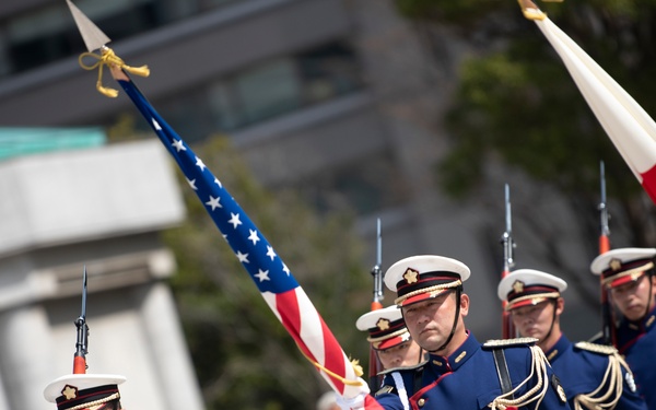 A military honor guard unit is marching in a formal ceremonial parade, carrying both the American flag and what appears to be the Japanese flag. The soldiers are dressed in distinctive navy blue and white dress uniforms with gold accents, white gloves, and white peaked caps adorned with insignia. The scene suggests a joint military ceremony, likely involving the United States and Japan.