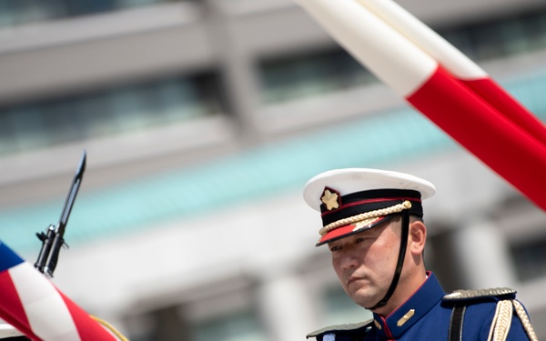 Two military personnel in formal dress uniforms are handling an American flag during what appears to be a ceremonial color guard presentation. Both individuals wear matching blue dress uniforms with gold epaulettes, white-topped caps with gold insignia, and white gloves. A second flag with red and white coloring is visible in the upper right corner of the frame.