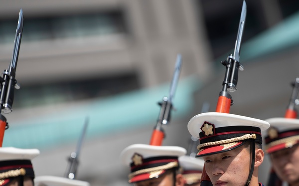A formation of military honor guard personnel marching in ceremonial dress uniforms. The soldiers are carrying rifles with fixed bayonets, suggesting a formal parade or ceremonial event. Their uniforms feature distinctive blue jackets with gold epaulettes and white peaked caps with red bands.