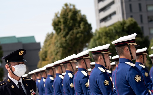 A formal military ceremony featuring a honor guard in dress uniforms standing in precise formation. A senior officer wearing a black uniform and face mask stands in the foreground, appearing to inspect or oversee the formation. The setting appears to be an outdoor plaza, likely in Japan based on the uniform insignia and architectural style.