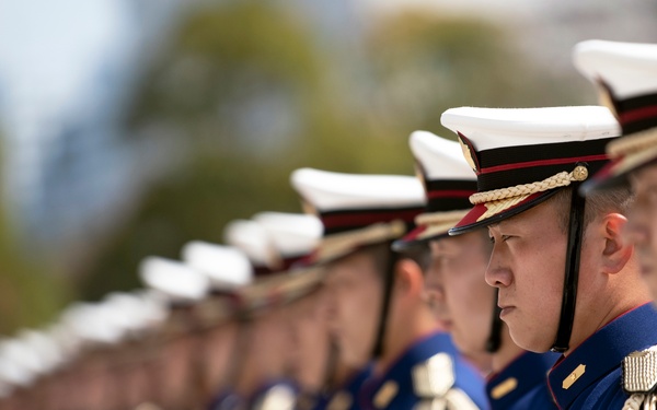 A formation of military honor guard personnel standing at attention in ceremonial dress uniforms. The soldiers are wearing dark navy blue jackets with gold epaulettes, aiguillettes, and brass buttons, paired with white-topped peaked caps with red and black bands. The image is captured with a shallow depth of field, keeping the foreground soldier sharp while the line recedes into blur.