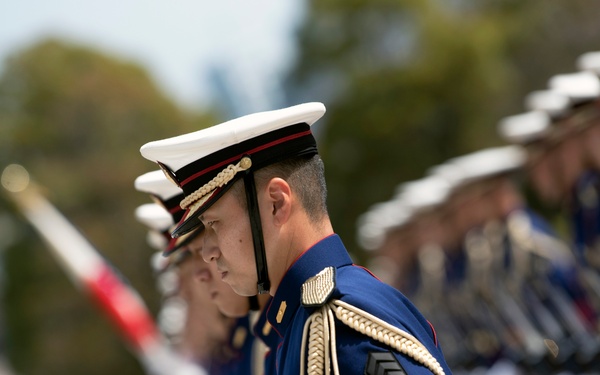 A formal military honor guard formation is shown standing at attention in dress uniforms. The soldiers wear distinctive blue ceremonial tunics with gold aiguillettes, white peaked caps with red bands, and carry rifles. The image uses shallow depth of field, with the foreground soldier in sharp focus and the formation receding into blur.