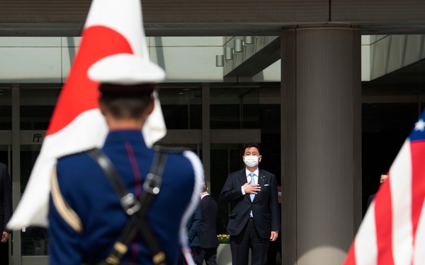 A formal military ceremony appears to be taking place outdoors, likely at a government building in Japan, involving officials from both Japan and the United States. Several dignitaries are standing at attention with hands over their hearts on raised platforms, suggesting a national anthem is being played. Both the Japanese and American flags are prominently displayed, and honor guard members in ceremonial uniforms are present.