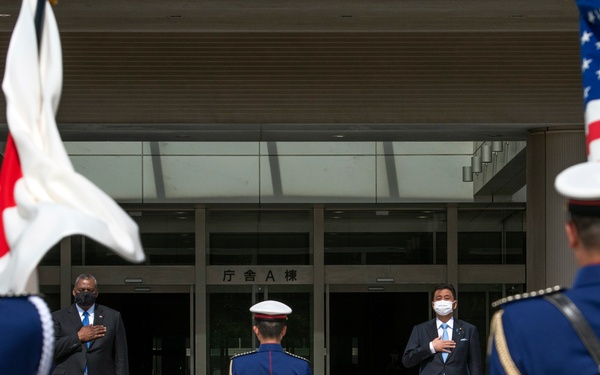 A formal military ceremony is taking place outside a Japanese government building, with two civilian officials standing at attention on raised platforms while honor guard members in dress uniforms hold flags. The presence of both the Japanese flag and the American flag suggests a bilateral diplomatic or defense meeting. Both civilian officials have their right hands placed over their hearts, indicating a national anthem ceremony.