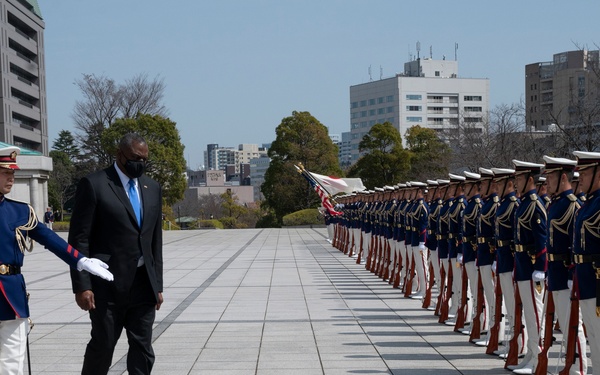A formal military honor guard ceremony is taking place at what appears to be a government or military facility in Japan. A civilian dignitary in a dark suit and face mask is being escorted by a Japanese military officer in ceremonial dress uniform past a long line of honor guard soldiers. Both American and Japanese flags are visible in the background, suggesting a bilateral diplomatic or military meeting.