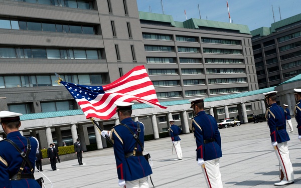 A military honor guard ceremony is taking place in an outdoor plaza, with personnel in formal dress uniforms carrying the American flag and what appears to be the Japanese flag. The soldiers are wearing distinctive blue tunics with white trousers and white caps, arranged in a formal line formation. The setting appears to be a government or military compound in Japan, based on the architectural style of the surrounding buildings.
