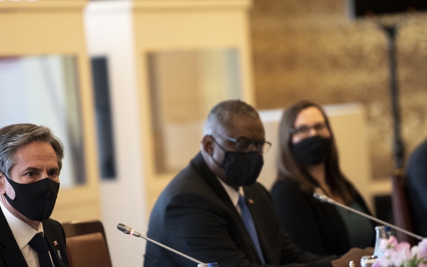 Four individuals are seated at a formal conference or diplomatic meeting table, all wearing black face masks. The setting appears to be an official high-level meeting, with microphones, water bottles, floral arrangements, and name placards visible on a navy blue tablecloth. The participants are dressed in formal business attire, suggesting a professional or governmental context.