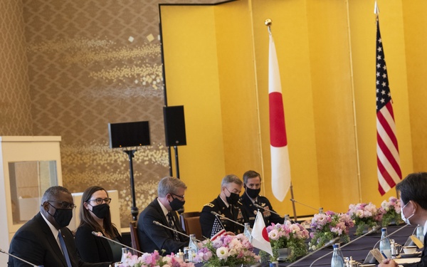 A formal bilateral diplomatic meeting is taking place between delegations seated on opposite sides of a long conference table. Japanese and American flags are prominently displayed in the background against a gold decorative screen, indicating a U.S.-Japan official meeting. Most participants are wearing black face masks, suggesting this occurred during the COVID-19 pandemic period.