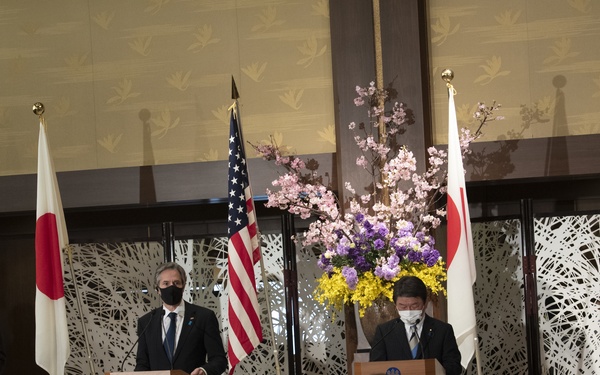 A formal diplomatic press conference or joint statement event featuring four officials standing at individual podiums. The setting displays alternating Japanese and American flags, suggesting a bilateral or joint US-Japan diplomatic meeting. A large floral arrangement with cherry blossoms and purple flowers serves as a centerpiece backdrop.