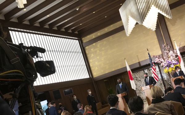 A formal press conference or diplomatic briefing is taking place in an elegant hall, with officials standing at podiums at the front of the room. Both Japanese and American flags are visible, indicating a bilateral diplomatic event. Members of the press are seated in rows with laptops, while a camera operator films from the left foreground.