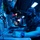 A military personnel member is seated at a combat information or operations console aboard what appears to be a naval vessel. The operator is wearing a headset, face mask, and a dark navy cap, working at a multi-screen tactical display station in a dimly lit, blue-toned environment. Additional crew members and equipment consoles are visible in the background.