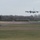 420th ABS supports Baltic Trident Exercise A large military or tanker aircraft is on final approach to land at an airfield, with landing gear deployed. The aircraft appears to be a gray, four-engine jet consistent with a KC-135 or similar tanker/transport type. The airfield is surrounded by bare winter trees and flat grassy terrain, suggesting a temperate climate location.