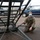 420th ABS supports Baltic Trident Exercise A military service member wearing olive drab/OCP camouflage uniform, a beanie, face mask, and communications headset is crouching near the base of a large metal maintenance stand or aircraft access ladder on what appears to be a flight line or airfield ramp. A U.S. Air Force aircraft (likely a KC-135 or similar tanker) is visible in the background along with military vehicles. The setting suggests an active airfield operations environment.