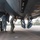420th ABS supports Baltic Trident Exercise Two military personnel in uniform are performing maintenance work on the underside of a large military aircraft on a flight line or tarmac. One individual is actively inspecting or working on a component beneath the fuselage while the other observes. The setting appears to be a military airfield, with visible ground support equipment and perimeter fencing in the background.