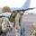 420th ABS supports Baltic Trident Exercise Three military personnel in OCP (Operational Camouflage Pattern) uniforms are standing on an aircraft flight line or tarmac. At least one individual is wearing a blue surgical face mask and a olive/tan beanie cap, suggesting this was taken during the COVID-19 pandemic era. A large military aircraft, likely a KC-135 or similar tanker/transport, is visible in the background.