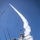 A missile is being launched from a naval warship, leaving a prominent white smoke trail ascending steeply into a clear blue sky. The low-angle photograph is taken from the deck of the vessel, emphasizing the scale of the superstructure and the dramatic trajectory of the projectile. A small flag and various radar/communications masts are visible on the ship's bridge structure.
