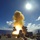 A naval warship is firing a missile from its deck in open ocean conditions. The launch produces a large plume of yellow-white smoke and a bright orange flame at the base of the missile. The image is captured from an onboard perspective under clear blue skies with bright sunlight.