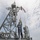 A naval crew member wearing a face mask and blue uniform is raising or handling signal flags on the mast structure of a military vessel. Multiple colorful signal flags are visible being hoisted on the rigging. The image is taken from a low angle, emphasizing the height of the ship's communications and mast superstructure against a partly cloudy sky.