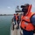 Three naval personnel are standing on the deck of a military patrol vessel in a harbor or port environment. The foreground sailor is wearing an orange life vest and a navy-branded face mask, suggesting this was taken during the COVID-19 pandemic period. Industrial port infrastructure and a large cargo or commercial vessel are visible in the background.
