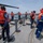A group of naval or coast guard personnel wearing orange life vests and blue uniforms are standing on the deck of a military or patrol vessel in a harbor. The crew members appear to be at their stations during a docking or maneuvering operation, with several large cargo ships visible in the background. Some personnel are wearing face masks, suggesting this was taken during the COVID-19 pandemic period.