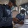 Two Ghana Navy personnel in blue uniforms and face masks are working together aboard what appears to be a naval vessel. The officer in the foreground, identified by a name tag reading 'FOLI' and wearing a cap marked 'GHANA NAVY', is referencing a device while a colleague writes in a logbook. The setting appears to be an interior cabin or bridge area of a ship.