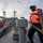 A naval or maritime personnel member wearing an orange life vest and face mask is handling a mooring rope on the deck of a military vessel in a harbor. Multiple naval ships are visible in the background docked at a port, with other crew members visible on adjacent vessels. The image appears to depict a docking or undocking operation during early morning or late afternoon light.